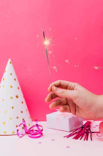 Kostenloses Foto nahaufnahme einer person, die wunderkerze vor partyhut hält; geschenkbox; luftschlangen vor rosa hintergrund