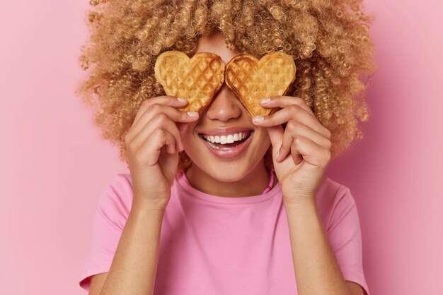 Nahaufnahme einer positiven Frau mit lockigen Haaren bedeckt die Augen mit herzförmigen hausgemachten Waffeln, die in einem lässigen T-Shirt gekleidet herumalbern, das über rosa Hintergrund isoliert ist Süßes Essen und Kalorien