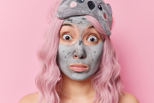 Nahaufnahme einer schockierten Frau, die eine nährende Schlammmaske zur Verjüngung trägt, die tägliche Schönheitsroutinen durchmacht, verwanzte Augen anstarrt hat lange gefärbte Haare einzeln auf rosafarbenem Hintergrund. Gesichtspflegekonzept