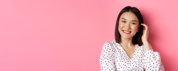 Kostenloses Foto nahaufnahme einer schönen asiatischen frau, die glücklich lächelt und einen neuen haarschnitt berührt, der über rosafarbenem hintergrund steht