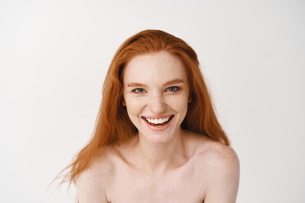 Nahaufnahme einer schönen blass rothaarigen Frau, die nackt auf einer weißen Wand posiert, glücklich lächelt und lacht und nackt auf der Studiowand steht
