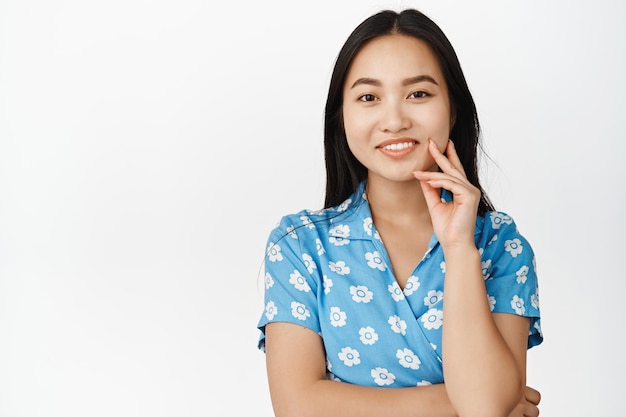 Nahaufnahme einer schönen brünetten asiatischen Frau, die selbstbewusst und interessiert in die Kamera schaut, die vor weißem Hintergrund steht