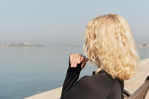 Nahaufnahme einer schönen erwachsenen blonden Frau, die auf einer Bank sitzt und auf das Meer und den blauen Himmel blickt Silhouette einer Frau Platz für Textidee für den Hintergrund