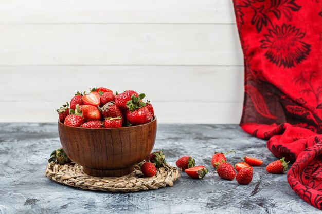 Nahaufnahme einer Schüssel Erdbeeren auf rundem Wicker-Tischset mit rotem Schal auf dunkelblauem Marmor und weißer Holzbrettoberfläche. horizontaler freier Speicherplatz für Ihren Text