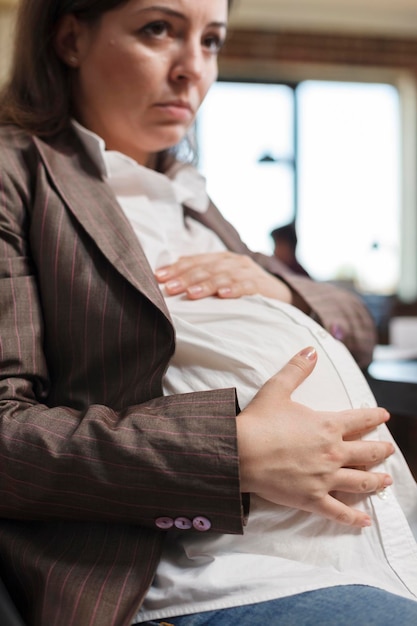 Nahaufnahme einer schwangeren Frau, die den Schwangerschaftsbauch berührt, während sie sich im Büroarbeitsplatz der Agentur befindet. Werdende Mutter sitzt am Arbeitsplatz der Marketingfirma und hält Babybauch.
