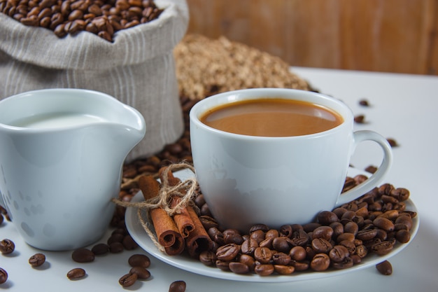 Nahaufnahme einer Tasse Kaffee mit Kaffeebohnen in einem Sack und einer Untertasse, Milch, trockenem Zimt auf Untersetzer und weißer Oberfläche. horizontal