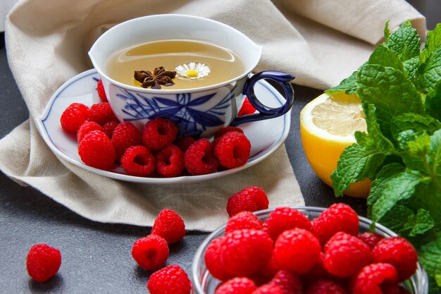 Nahaufnahme einer Tasse Kamillentee mit Zitrone, Minzblättern, Himbeeren in Untertasse auf Stoff