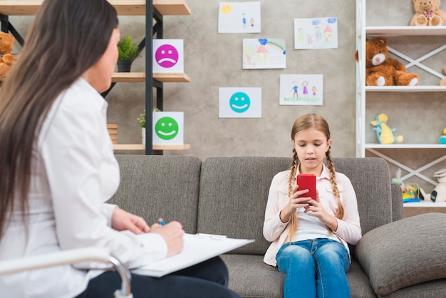 Nahaufnahme einer weiblichen Psychologeschreibensanmerkung über Klemmbrett vor dem Mädchen, das Handy verwendet