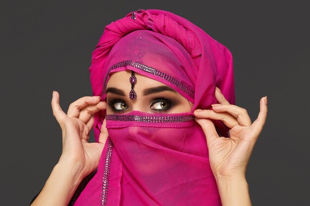 Nahaufnahme einer wunderschönen jungen Frau mit ausdrucksstarken rauchigen Augen, die den schicken rosa Hijab trägt, der mit Pailletten und Schmuck verziert ist. Sie hält den Schal mit ihren Händen und schaut weg auf eine Dunkelheit