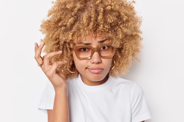 Nahaufnahme einer zögernden Frau, die die Hand hebt, die Lippen hat, hat zweifelhaften Ausdruck, Gesichter, schwierige Wahl, trägt eine Brille, lässiges T-Shirt, das über weißem Hintergrund isoliert ist. Konzept der menschlichen Reaktionen