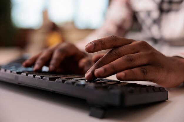 Nahaufnahme eines afroamerikanischen männlichen Inhaltserstellers, der Post für soziale Medien auf der Computertastatur im heimischen Wohnzimmer schreibt. Selektiver Fokus auf Männerhände, die beim Remote-Tipp-Blog-Artikel arbeiten.