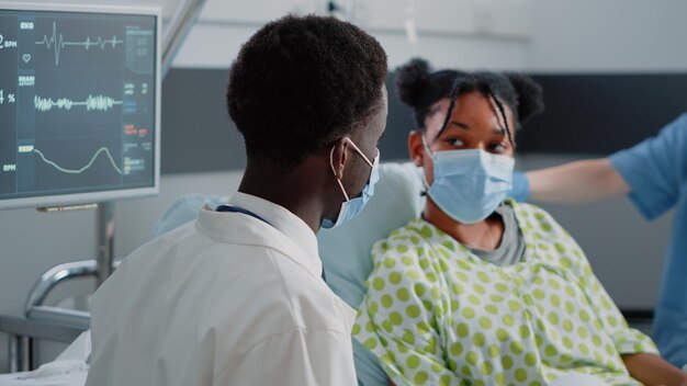 Nahaufnahme eines Arztes, der mit einem jungen Patienten im Bett spricht und Gesichtsmasken trägt. Mann, der als Spezialist arbeitet und während der Coronavirus-Pandemie mit kranken Frauen über Gesundheitsversorgung und Medizin spricht