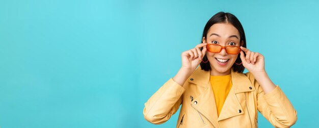 Nahaufnahme eines asiatischen Mädchens mit Sonnenbrille, das glücklich und überrascht auf dem Gesicht vor blauem Hintergrund aussieht