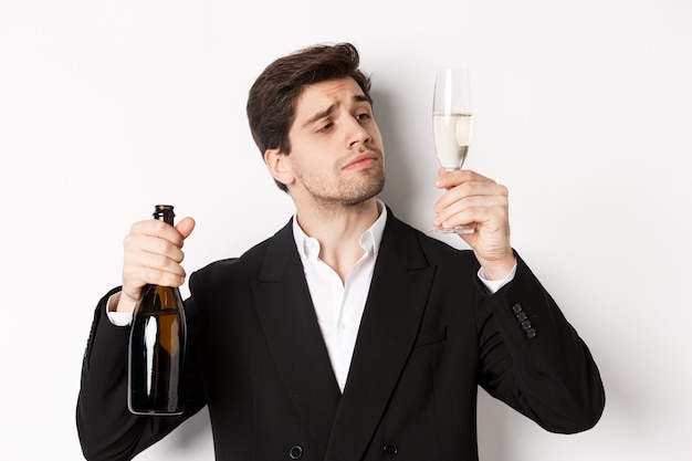 Nahaufnahme eines attraktiven Mannes im trendigen Anzug, der Champagner probiert, Glas betrachtet und vor weißem Hintergrund steht.