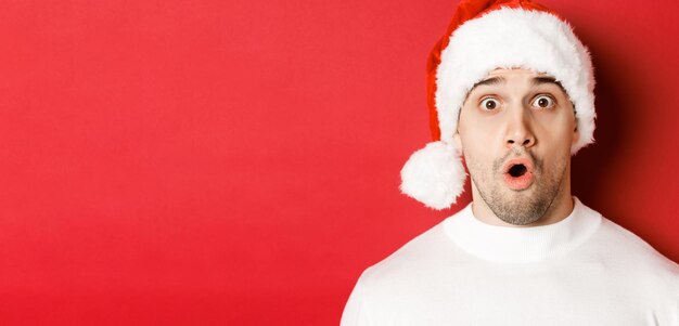 Nahaufnahme eines attraktiven überraschten Mannes in Weihnachtsmütze, der Wow sagt und etwas Interessantes betrachtet, das vor rotem Hintergrund steht.