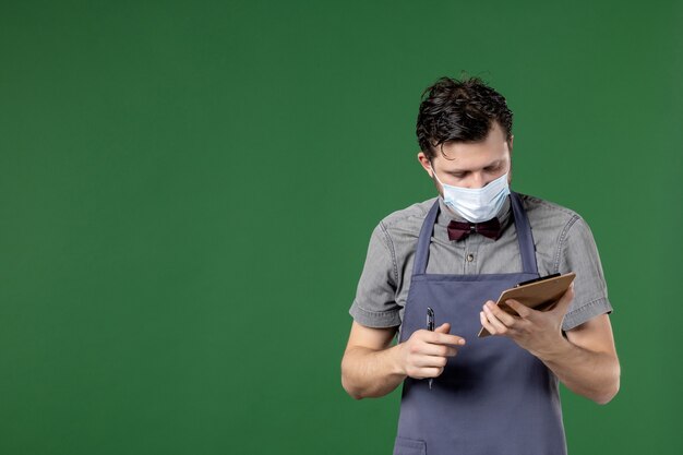 Nahaufnahme eines beschäftigten männlichen Kellners in Uniform mit medizinischer Maske und Auftragsbuch auf grünem Hintergrund