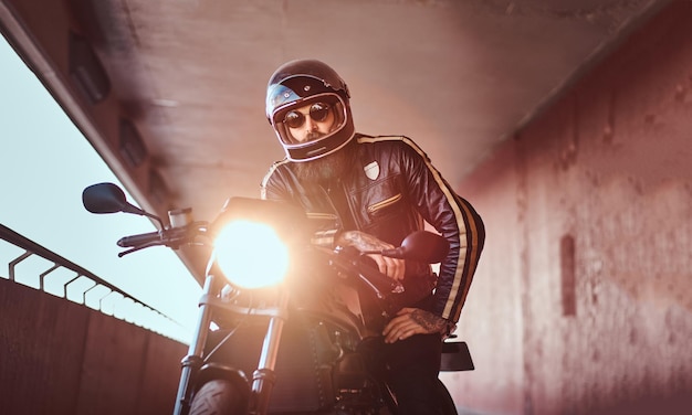 Nahaufnahme eines brutalen bärtigen Bikers mit Helm und Sonnenbrille in einer schwarzen Lederjacke, der auf einem Retro-Motorrad mit eingebautem Scheinwerfer sitzt.