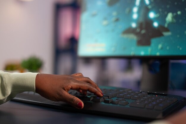 Nahaufnahme eines erfolgreichen Profispielers, der während der Online-Meisterschaft Computertastatur drückt, während er Weltraum-Shooter-Videospiele spielt. Spielerin sitzt am Schreibtisch im Gaming-Studio mit RGB-Geräten