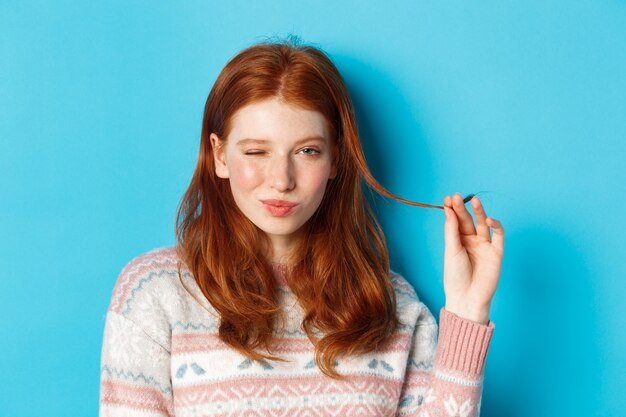 Nahaufnahme eines frechen rothaarigen Mädchens, das mit einer Haarsträhne spielt, in die Kamera zwinkert und lächelt und auf blauem Hintergrund steht