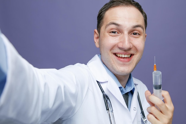 Nahaufnahme eines fröhlichen jungen männlichen Arztes, der einen medizinischen Bademantel und ein Stethoskop um den Hals trägt und in die Kamera blickt, die die Hand in Richtung Kamera ausstreckt und eine Spritze mit Nadel zeigt, die ein Selfie macht