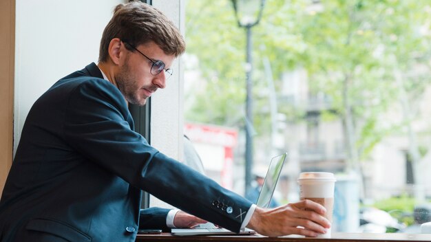 Nahaufnahme eines Geschäftsmannes unter Verwendung des Laptops, der Wegwerfkaffeetasse im Café hält
