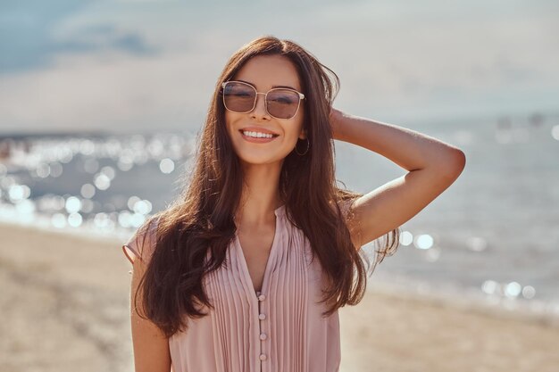 Nahaufnahme eines glücklichen, schönen, brünetten Mädchens mit langen Haaren, Sonnenbrille und Kleid am Strand.