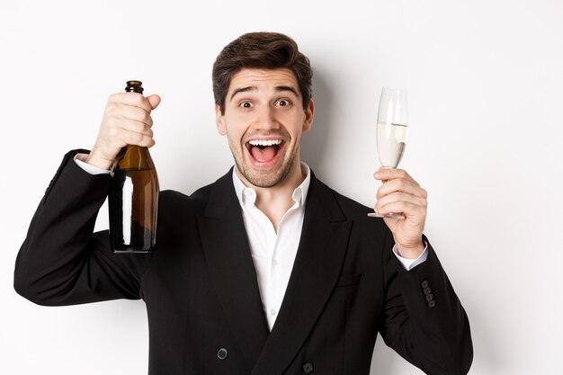 Nahaufnahme eines gutaussehenden lächelnden Mannes im schwarzen Anzug, der einen Toast macht, Champagner und Glas hält, das neue Jahr feiert und vor weißem Hintergrund steht