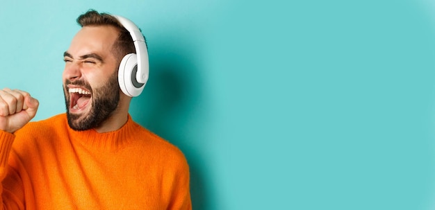 Kostenloses Foto nahaufnahme eines gutaussehenden modernen mannes, der musik in kopfhörern hört, die in einem orangefarbenen pullover über türkis stehen