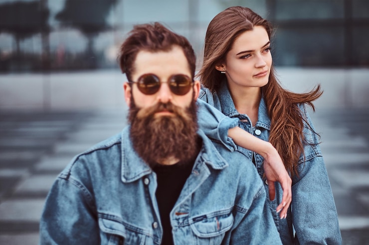 Kostenlose Foto nahaufnahme eines hipster-paares eines brutalen bärtigen mannes mit sonnenbrille und seiner freundin in jeansjacken vor einem wolkenkratzer.