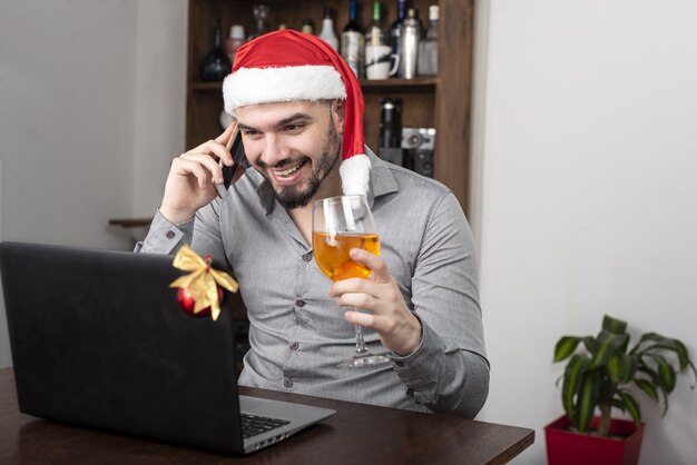 Nahaufnahme eines hispanischen Mannes, der eine Weihnachtsmütze trägt, seinen Wein genießt und am Telefon spricht
