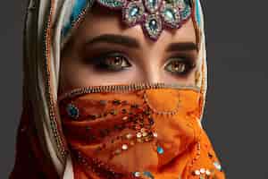Kostenloses Foto nahaufnahme eines hübschen mädchens mit professionellem make-up, das einen farbenfrohen hijab trägt, der mit pailletten und schmuck verziert ist. sie posiert im studio und schaut auf einem dunklen hintergrund weg. menschliche gefühle,