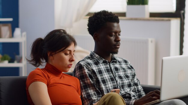 Nahaufnahme eines interracial Paares mit modernen Geräten zu Hause. Junge multiethnische Ehemänner und -frauen, die Smartphone und Laptop benutzen, um sich im Wohnzimmer zu entspannen und lustige Aktivitäten zu machen?