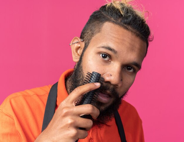 Nahaufnahme eines jungen afroamerikanischen männlichen Friseurs in Uniform, der seinen Schnurrbart kämmt