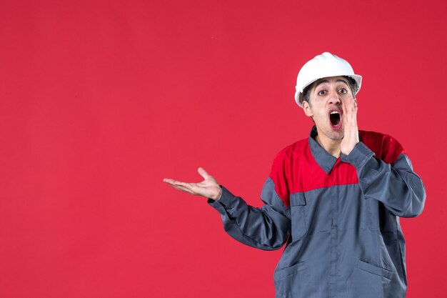 Nahaufnahme eines jungen Arbeiters in Uniform mit Schutzhelm, der auf etwas auf der rechten Seite zeigt, der jemanden an einer isolierten roten Wand anruft?