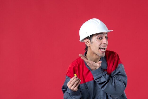 Nahaufnahme eines jungen Arbeiters in Uniform mit Schutzhelm und Abnehmen eines Ohrstöpsels an einer isolierten roten Wand