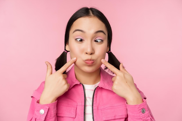 Nahaufnahme eines jungen asiatischen Mädchens, das ihre Grübchen zeigt, die dumme Wangen stoßen und lustige Gesichter machen, die über rosa Hintergrund stehen