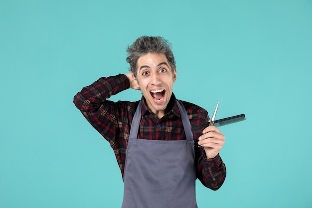 Nahaufnahme eines jungen lächelnden, glücklichen männlichen friseurs, der eine graue schürze trägt und einen scherenkamm auf einer pastellblauen oberfläche hält