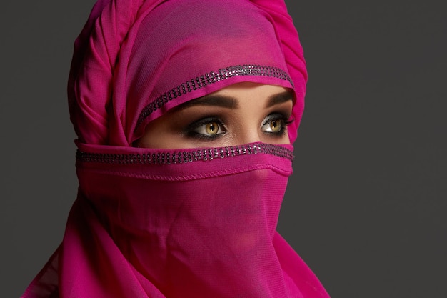 Nahaufnahme eines jungen Mädchens mit hellem Make-up, das den rosa Hijab trägt, der mit Pailletten verziert ist. Sie posiert im Studio und schaut auf einem dunklen Hintergrund weg. Menschliche Emotionen, Gesichtsausdruck