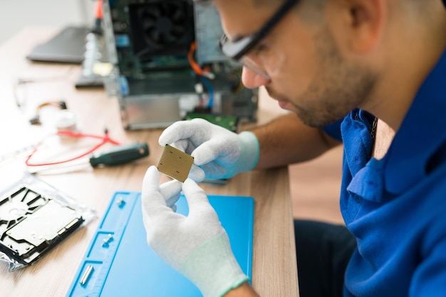 Nahaufnahme eines jungen Technikers, der eine Schutzbrille und Handschuhe trägt, während er den beschädigten Mikroprozessorchip eines Computers überprüft