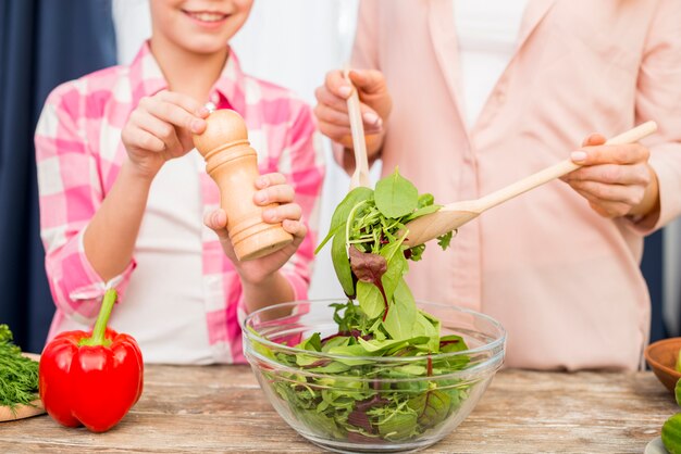 Nahaufnahme eines mahlenden Pfeffers des Mädchens in die Salatschüssel vorbereitet von ihrer Mutter