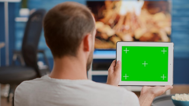 Nahaufnahme eines Mannes, der ein digitales Tablet mit grünem Bildschirm hält und sich ein Webinar auf dem Sofa vor dem Fernseher ansieht. Person, die sich ein Touchscreen-Gerät mit Chroma-Key ansieht und zu Hause an einem Online-Kurs teilnimmt.