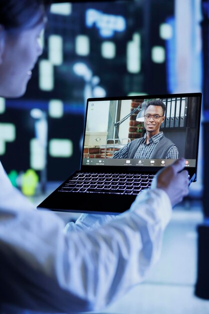 Nahaufnahme eines Mannes im Videogespräch mit einem Kollegen, während er nachts durch die Straßen der Stadt spaziert und ihm Arbeitsaktualisierungen gibt. Person nutzt Laptop, um das Stadtbild des Nachtlebens eines Kollegen vor verschwommenem Hintergrund zu zeigen