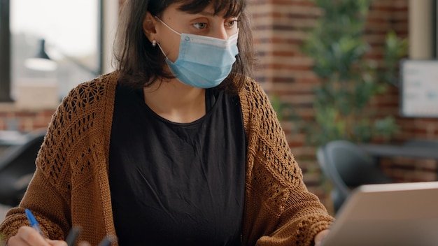 Nahaufnahme eines Mitarbeiters, der mit einem Laptop arbeitet und sich Notizen auf Papieren mit Diagrammen macht und eine Gesichtsmaske trägt. Frau, die Computer verwendet, um während der Coronavirus-Pandemie an Geschäftsprojekten zu arbeiten.