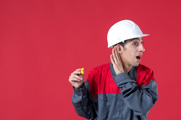 Nahaufnahme eines neugierigen jungen Baumeisters in Uniform mit Schutzhelm und Ohrstöpseln, die dem letzten Tratsch an einer isolierten roten Wand zuhören