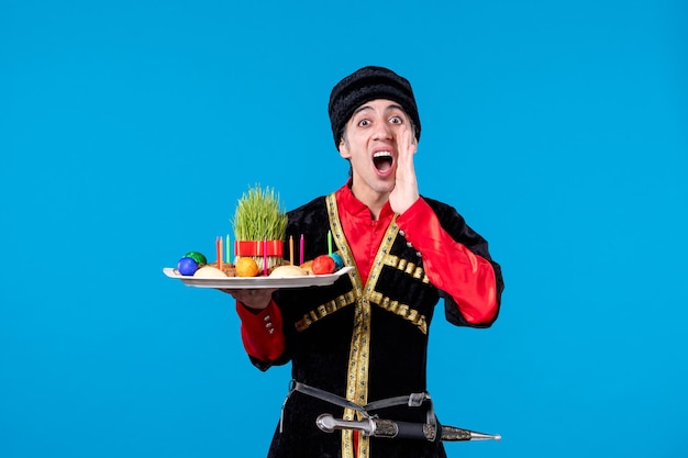 Nahaufnahme eines neugierigen Mannes in traditioneller Kleidung, der ein Tablett mit nationalen Süßwaren hält, das jemanden auf blauem Hintergrund anruft