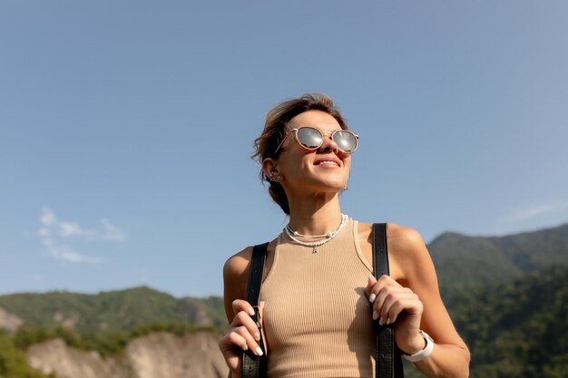 Nahaufnahme eines Outdoor-Porträts einer attraktiven hübschen Frau mit Sonnenbrille und Rucksack, die an einem sonnigen, warmen Tag zwischen Bergen unterwegs ist