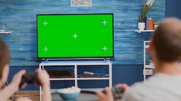 Nahaufnahme eines Paares, das Controller hält, die Action-Konsolenspiele auf Greenscreen-Fernsehern spielen, auf der Couch sitzen und Popcorn essen. Junge Gamer verbringen ihre Freizeit mit Online-Spielen auf Chroma-Key-Mockup-Displays.