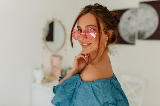 Nahaufnahme eines Porträts einer stilvollen, charmanten Frau mit wunderbarem Lächeln, die eine runde Brille und ein blaues T-Shirt trägt und vor hellem Hintergrund in die Kamera lächelt