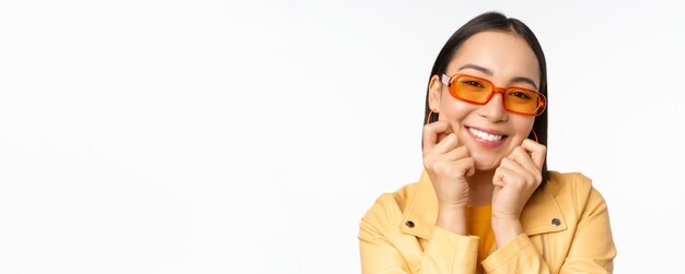 Nahaufnahme eines Porträts einer trendigen asiatischen Frau mit Sonnenbrille, die ihr Gesicht berührt und romantisch in die Kamera lächelt, die über weißem Hintergrund steht