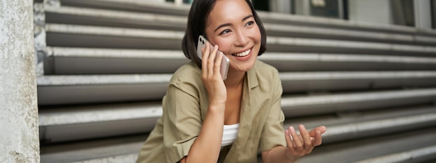 Nahaufnahme eines Porträts eines lächelnden asiatischen Mädchens, das auf dem Handy spricht und jung draußen auf einer Straßentreppe sitzt